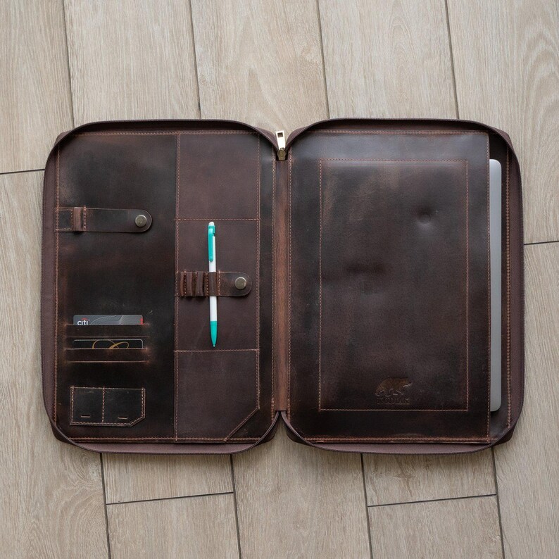 May include: A dark brown leather portfolio case featuring a full-length zipper, multiple interior card slots, a pen holder with a white and teal pen, and a dedicated sleeve containing a laptop. A small bear logo is embossed on the interior.