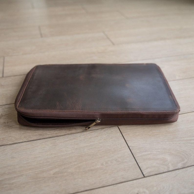 May include: A rectangular dark brown leather laptop sleeve with a smooth, matte finish and visible natural grain. The case features a matching brown fabric border and a metal zipper closure that is partially open along the front edge.