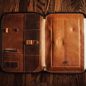 May include: A brown leather portfolio case with a zipper closure, featuring multiple interior pockets for cards, pens, and documents, and a dedicated sleeve for a laptop. The interior is embossed with a bear logo and the text KODIAK.