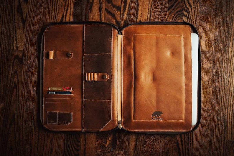 May include: A brown leather portfolio case with a zipper closure, featuring multiple interior pockets for cards, pens, and documents, and a dedicated sleeve for a laptop. The interior is embossed with a bear logo and the text KODIAK.
