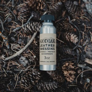 May include: A silver bottle with a brown label that reads "Kodiak Leather Dressing", "Restores | Protects | Waterproofs", and "3oz". The bottle is sitting on a bed of pine needles and cones.