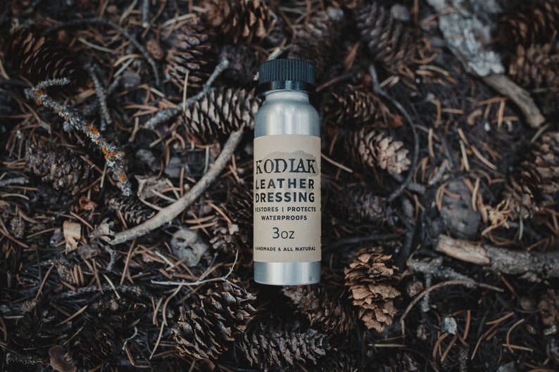 May include: A silver bottle with a brown label that reads "Kodiak Leather Dressing", "Restores | Protects | Waterproofs", and "3oz". The bottle is sitting on a bed of pine needles and cones.