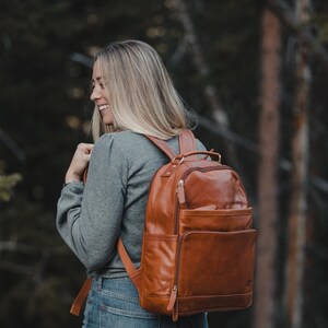 May include: A brown leather backpack with multiple pockets and a top handle. The backpack is worn by a person in a gray shirt and blue jeans, with a forest background.