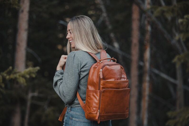 May include: A brown leather backpack with multiple pockets and a top handle. The backpack is worn by a person in a gray shirt and blue jeans, with a forest background.