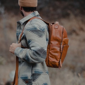 May include: A brown leather backpack with multiple pockets and a zipper closure. The backpack is carried over the shoulder with brown leather straps. The person is wearing a patterned jacket with blue and gray accents.