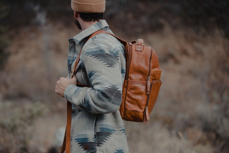 May include: A brown leather backpack with multiple pockets and a zipper closure. The backpack is carried over the shoulder with brown leather straps. The person is wearing a patterned jacket with blue and gray accents.