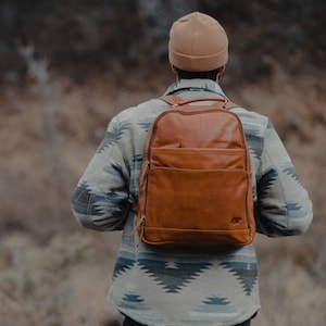 May include: A brown leather backpack with a front pocket and top handle is worn by a person. The backpack has a bear logo on the bottom right. The person is wearing a patterned jacket and a tan beanie.