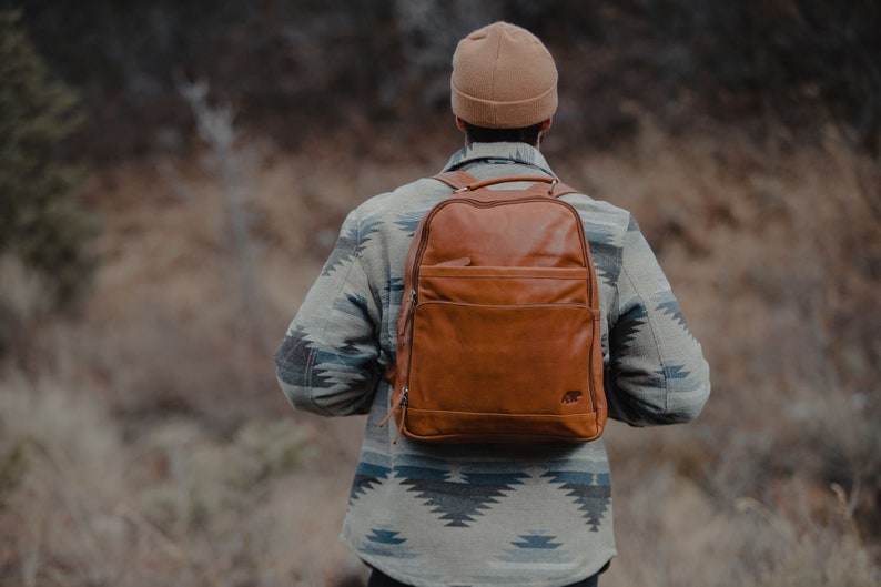 May include: A brown leather backpack with a front pocket and top handle is worn by a person. The backpack has a bear logo on the bottom right. The person is wearing a patterned jacket and a tan beanie.