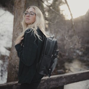 May include: A black leather backpack with multiple compartments and silver-toned hardware. The backpack is worn by a person in a dark green jacket, standing on a wooden bridge in a snowy outdoor setting.