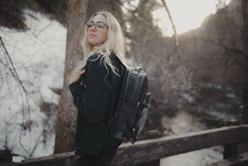 May include: A black leather backpack with multiple compartments and silver-toned hardware. The backpack is worn by a person in a dark green jacket, standing on a wooden bridge in a snowy outdoor setting.