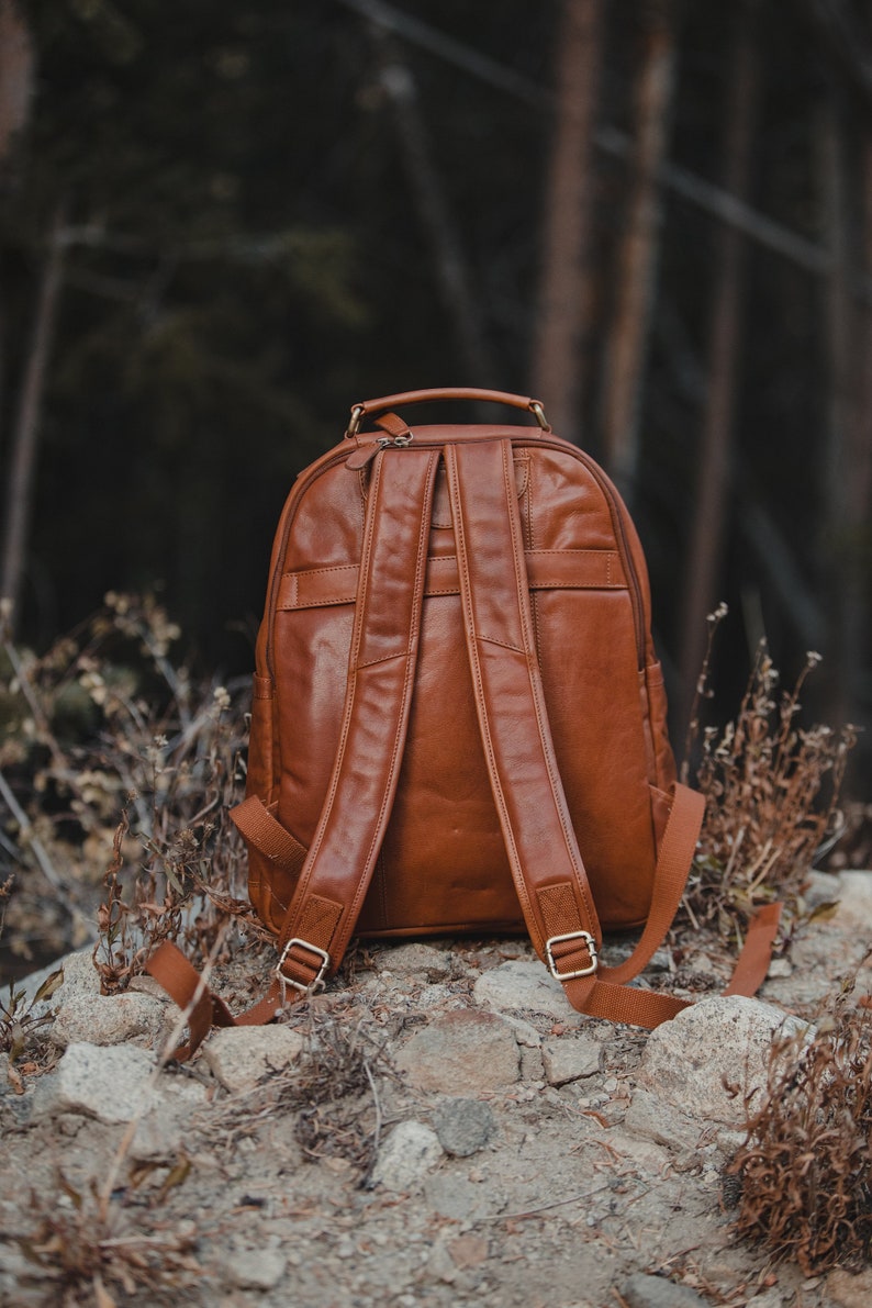 May include: A brown leather backpack with adjustable shoulder straps and a top handle. The backpack has a zippered main compartment and a front pocket. The straps are secured with gold-colored metal buckles. The backpack is sitting on a rocky surface.