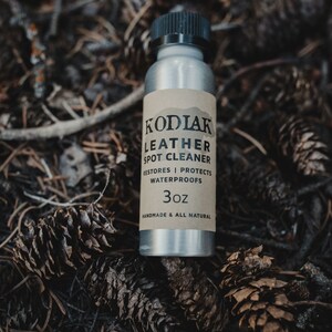 May include: A silver bottle with a black cap and a brown label that reads "Kodiak Leather Spot Cleaner", "Restores | Protects Water Proofs", and "3oz Handmade & All Natural". The bottle is sitting on a bed of pine needles and cones.