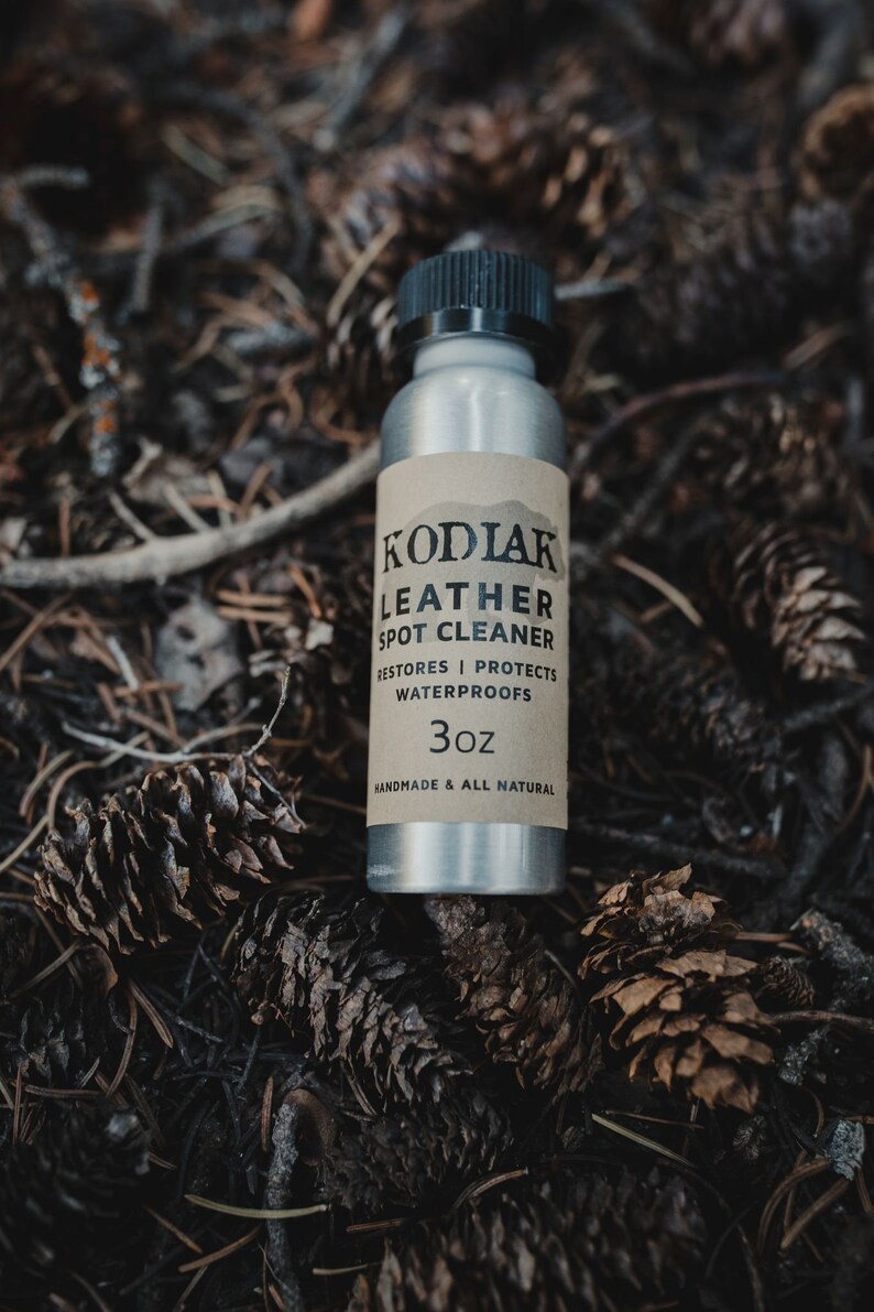 May include: A silver bottle with a black cap and a brown label that reads "Kodiak Leather Spot Cleaner", "Restores | Protects Water Proofs", and "3oz Handmade & All Natural". The bottle is sitting on a bed of pine needles and cones.