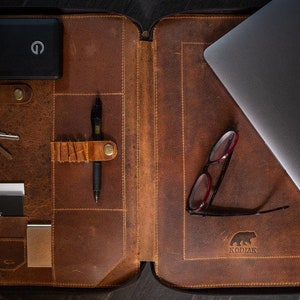 May include: A brown leather portfolio case featuring an interior organizer with slots for a black power bank, keys, a pen, business cards, and a laptop. The interior is embossed with a bear logo and the text KODIAK, with visible stitching.