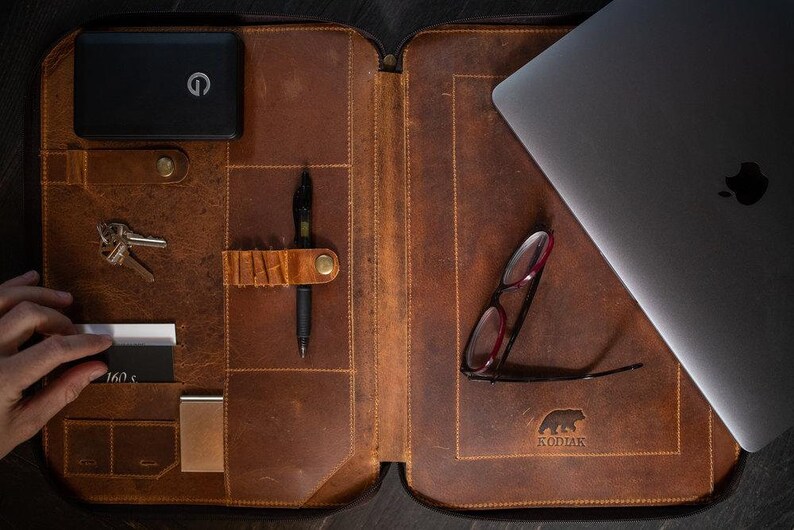May include: A brown leather portfolio case featuring an interior organizer with slots for a black power bank, keys, a pen, business cards, and a laptop. The interior is embossed with a bear logo and the text KODIAK, with visible stitching.