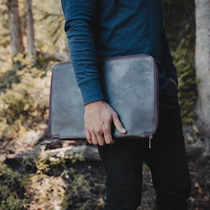 May include: A rectangular dark gray leather laptop sleeve with a smooth finish and a dark brown zipper closure. The case features a slim profile and is held horizontally, showcasing a clean, minimalist design with visible stitching along the edges.