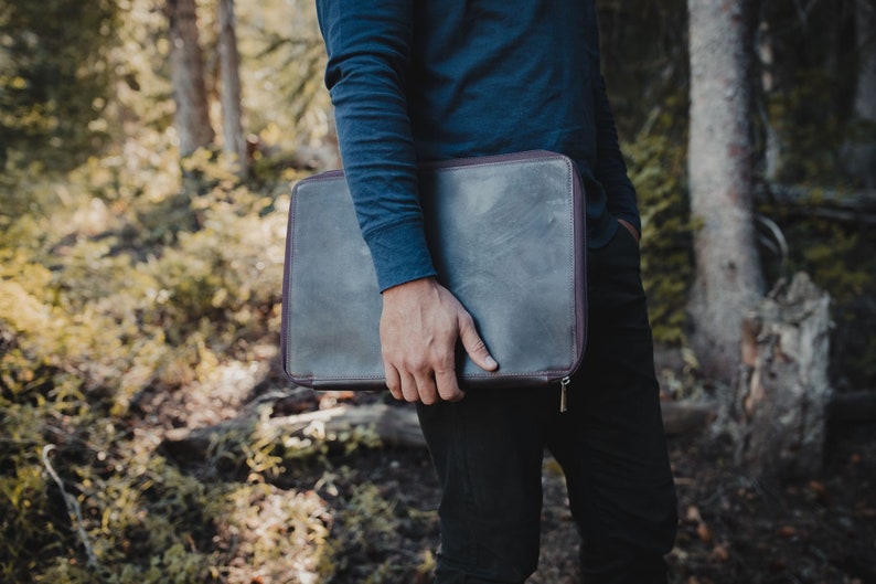 May include: A rectangular dark gray leather laptop sleeve with a smooth finish and a dark brown zipper closure. The case features a slim profile and is held horizontally, showcasing a clean, minimalist design with visible stitching along the edges.