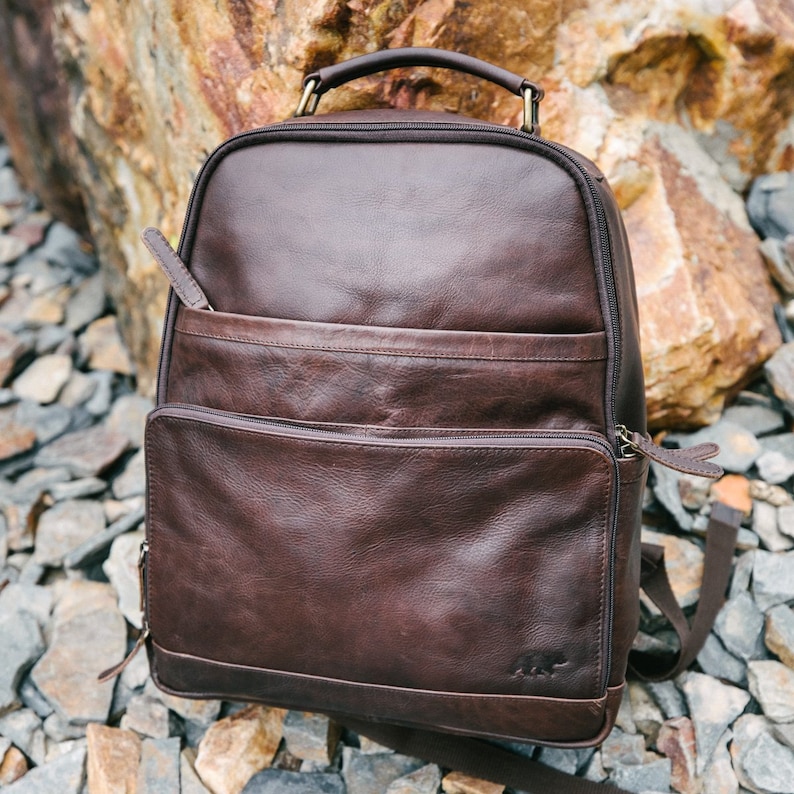 May include: A brown leather backpack with multiple zippered compartments and a top handle. The backpack has a front pocket and a small logo on the bottom. The bag is made of a rich, dark brown leather.