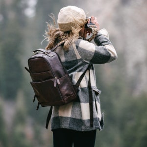 May include: A dark brown leather backpack with multiple pockets and a top handle. The backpack is worn by a person wearing a beige beanie, a plaid jacket in shades of gray and white, and black pants. The person is holding a camera.