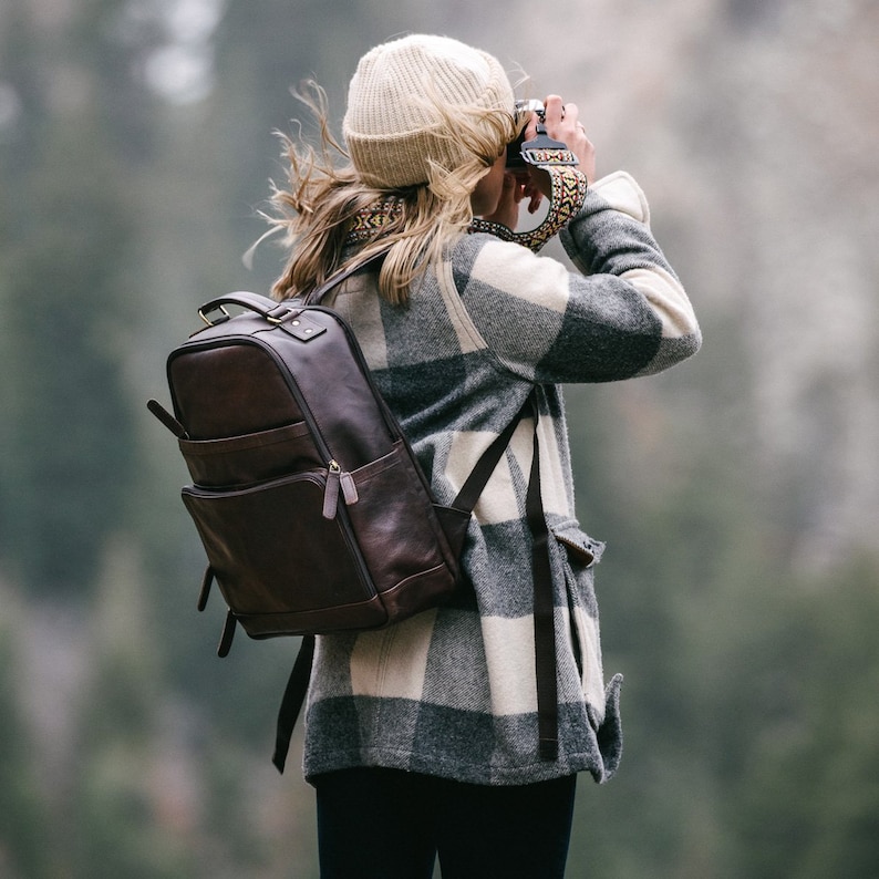 May include: A dark brown leather backpack with multiple pockets and a top handle. The backpack is worn by a person wearing a beige beanie, a plaid jacket in shades of gray and white, and black pants. The person is holding a camera.
