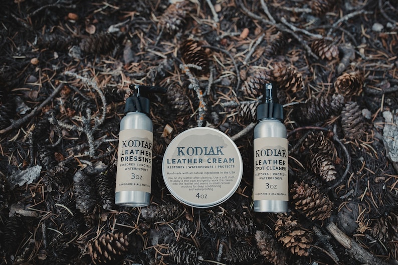 May include: Three silver bottles with black labels and white text. The bottles contain leather care products: Leather Dressing, Leather Cream, and Leather Spot Cleaner. Each bottle is labeled with the brand name "Kodiak" and the size in ounces. The bottles are arranged on a bed of pine needles and cones.