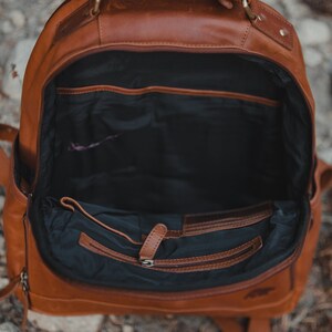 May include: Open brown leather backpack with multiple interior pockets and a zippered compartment. The bag has a top handle and adjustable shoulder straps. The interior is black.