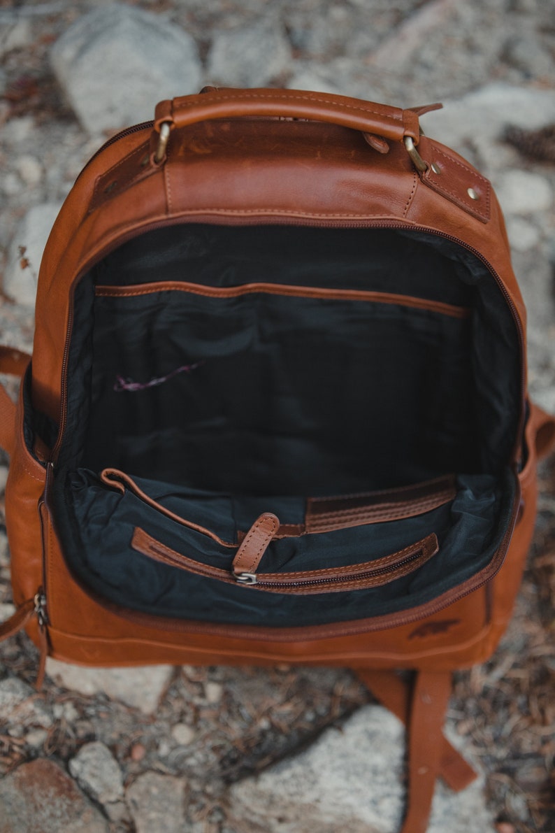 May include: Open brown leather backpack with multiple interior pockets and a zippered compartment. The bag has a top handle and adjustable shoulder straps. The interior is black.