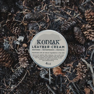 May include: A white tin container with a brown bear illustration and the text "KODIAK LEATHER CREAM RESTORES WATERPROOFS PROTECTS Handmade with all natural ingredients in the USA Use on dry leather after cleaning. Use a soft cloth to apply a thin coat and gently work the cream into the leather and seams in small circular motions for deep conditioning and waterproofing 4oz".