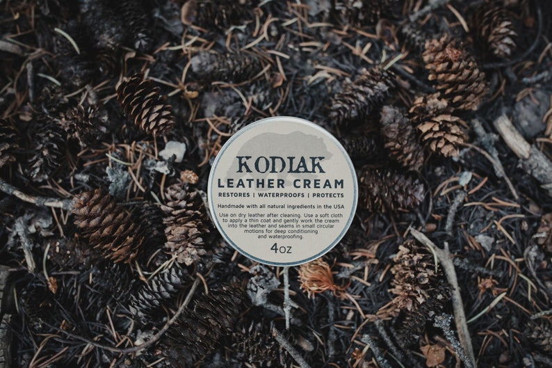 May include: A white tin container with a brown bear illustration and the text "KODIAK LEATHER CREAM RESTORES WATERPROOFS PROTECTS Handmade with all natural ingredients in the USA Use on dry leather after cleaning. Use a soft cloth to apply a thin coat and gently work the cream into the leather and seams in small circular motions for deep conditioning and waterproofing 4oz".