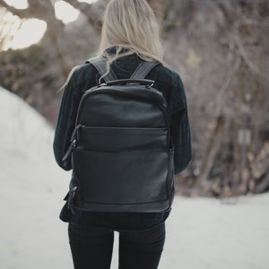 May include: A black leather backpack with multiple compartments and a top handle is worn by a person walking in a snowy outdoor setting. The backpack has a sleek, modern design with visible zippers and a front pocket.