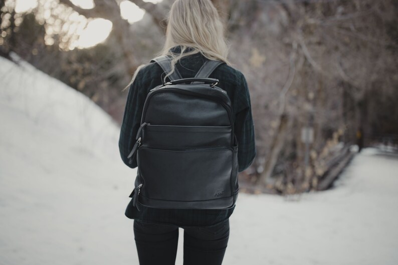 May include: A black leather backpack with multiple compartments and a top handle is worn by a person walking in a snowy outdoor setting. The backpack has a sleek, modern design with visible zippers and a front pocket.