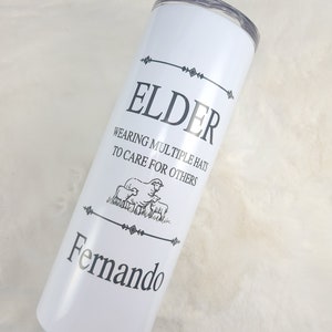 Puede incluir: Un vaso blanco de acero inoxidable con texto negro que dice "Elder Wearing Multiple Hats To Care For Others" y "Fernando".