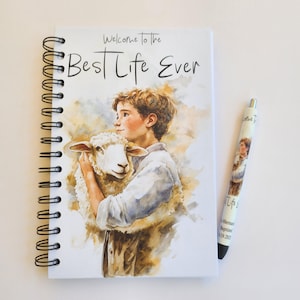 May include: A white spiral bound journal with the text "Welcome to the Best Life Ever" on the cover. The cover also features a watercolor illustration of a young boy holding a white lamb. A white pen with a matching illustration is included.