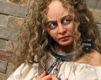 May include: A woman with long, curly brown hair is wearing a white shirt and a pair of metal shackles around her neck and wrists. The shackles are connected by a chain. She is looking down at her hands.