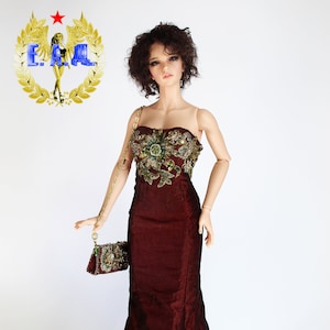 May include: A doll wearing a long, burgundy gown with gold and green embellishments. The gown has a fitted bodice and a flowing skirt. The doll is holding a small gold purse.