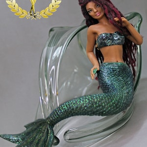 May include: A mermaid figurine with long red dreadlocks, a green and blue iridescent tail, and a black and silver bikini top. The figurine is posed sitting in a clear glass bowl.