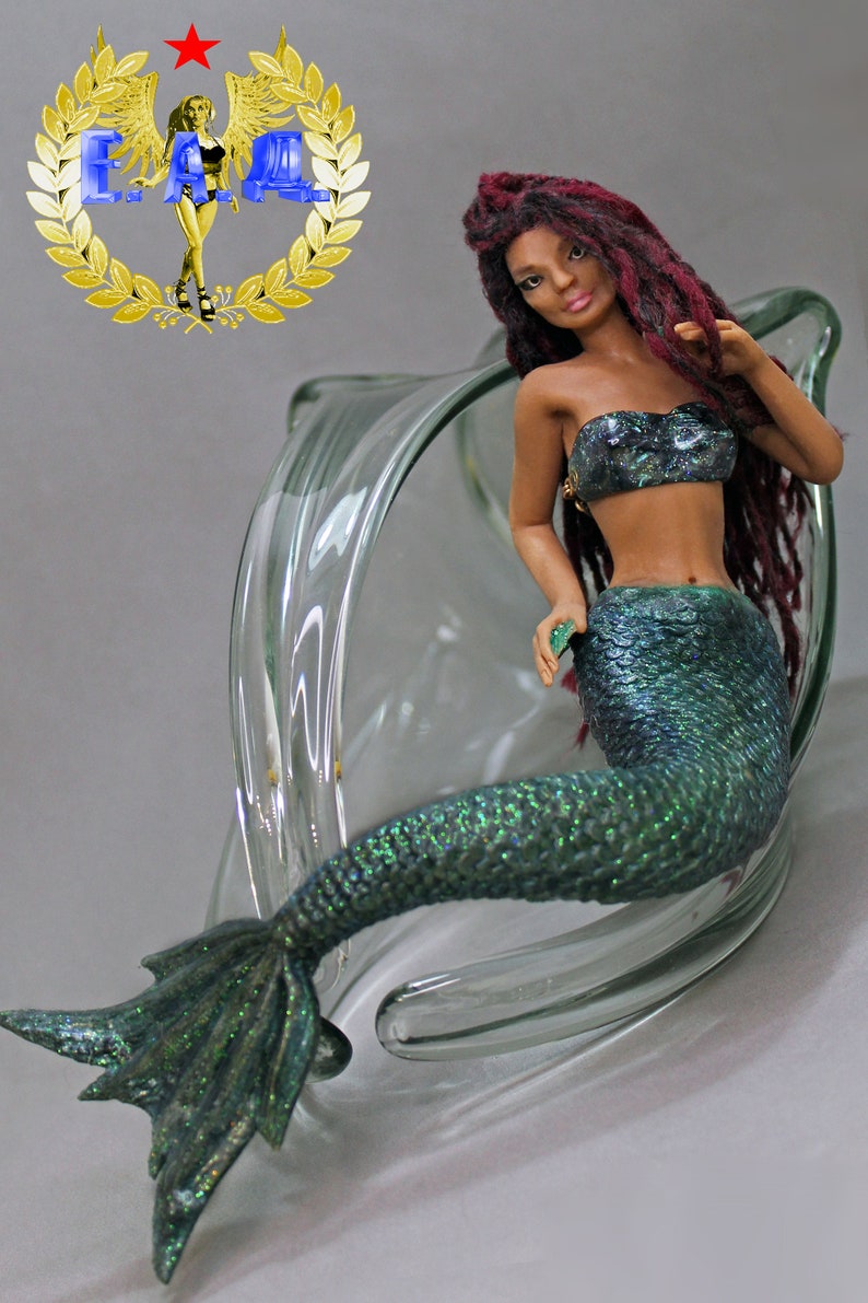 May include: A mermaid figurine with long red dreadlocks, a green and blue iridescent tail, and a black and silver bikini top. The figurine is posed sitting in a clear glass bowl.