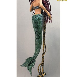 May include: A green mermaid figurine with a long, red, braided wig. The mermaid is standing on a gold, vine-like stand with a base decorated with green leaves and gems.