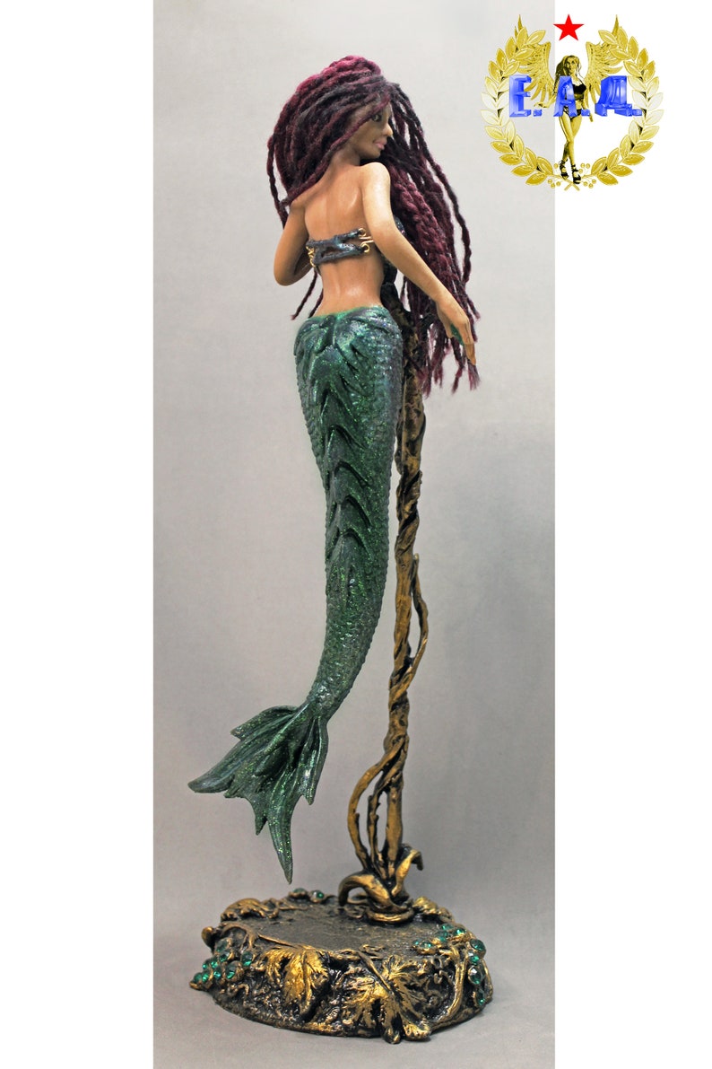 May include: A green mermaid figurine with a long, red, braided wig. The mermaid is standing on a gold, vine-like stand with a base decorated with green leaves and gems.
