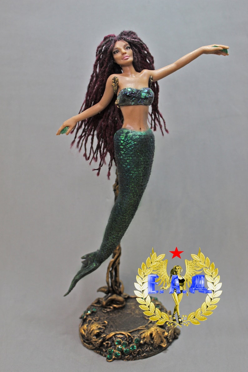May include: A green and iridescent mermaid figurine with long, dark red dreadlocks. The mermaid is holding a green gemstone in each hand. The figurine is standing on a gold-colored base with green gemstones.