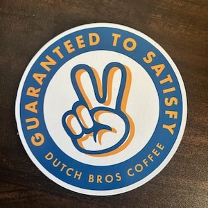 May include: A white round sticker with a blue circle around the edge. The circle has the text "Guaranteed to Satisfy" in yellow letters. Inside the circle is a peace sign with an orange outline. Below the circle is the text "Dutch Bros Coffee" in blue letters.