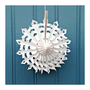 May include: A white paper snowflake decoration hanging from a blue wall. The snowflake is made of folded paper and has a delicate, intricate design.
