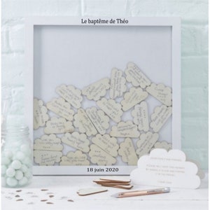 Peut inclure: Des cartes en bois en forme de nuage blanches avec des messages écrits à la main sont affichées dans un cadre blanc. Le cadre est intitulé "Le baptême de Théo" et la date "18 juin 2020" est écrite sous le cadre. Une carte en forme de nuage blanche avec un message est sur la table sous le cadre.