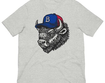 May include: A light grey t-shirt with a black and white illustration of a bison wearing a blue and red baseball cap. The cap has a white "B" on the front. The bison's fur and facial features are highly detailed.