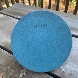 May include: A circular blue polishing pad with a textured surface and the number 600 printed in black near the top center. The pad is mounted on a clear plastic stand, displaying a consistent grit texture across its entire flat face.