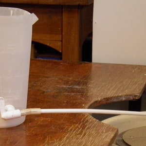 Könnte beinhalten: Eine weiße Plastikkanne mit einem Ausguss und einem weißen Plastikrohr, das an den Ausguss angeschlossen ist. Die Kanne steht auf einem Holztisch.