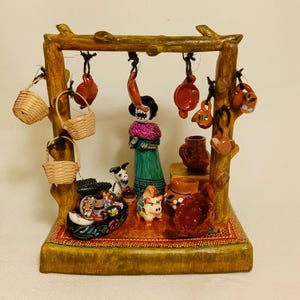 Pode incluir: Uma barraca de mercado em miniatura com tema Dia dos Mortos com uma figura esquelética, um porco e um cão. A barraca é feita de madeira e tem uma variedade de pequenos vasos de barro e cestas penduradas.