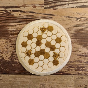 May include: A white round patch with a gold embroidered honeycomb design and three bees.
