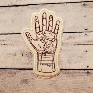 May include: A beige fabric patch with a brown embroidered image of a hand with lines and numbers, depicting a palmistry chart.