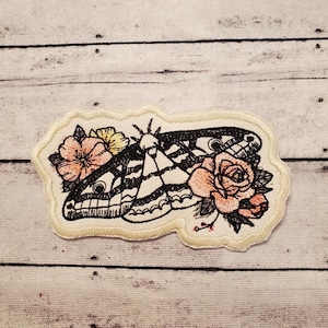May include: Embroidered iron-on patch featuring a black and white moth with pink and yellow flowers. The patch is on a white background.